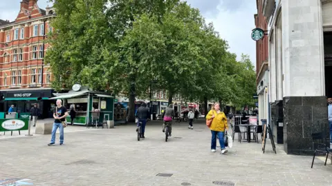 Emma Baugh/BBC A street with people walking and cycling, and trees in full leaf.
