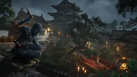 Ubisoft A still from the Assassins Creed game, showing a character dressed in blue covertly overlooking an area with trees and a home - looking at another male character who is standing on a bridge below.