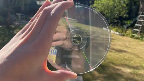 Max Gowers A hand holds a transparent CD in front of the camera. In the background, the green grass of a garden is visible, and can also be seen through the CD itself.