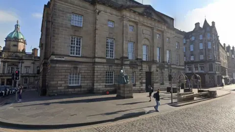 The sandstone court building is bathed in sunlight. There are cobbles on the road in the foreground and a man crossing the road.