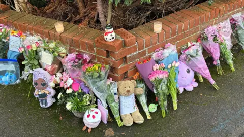 Bunches of brightly flowers line a street corner, around a low brick wall. Some of the bunches of flowers have handwritten notes on them, and there are also several stuffed toys. On the wall are several dandles, including a candle holder shaped like a snowman. 