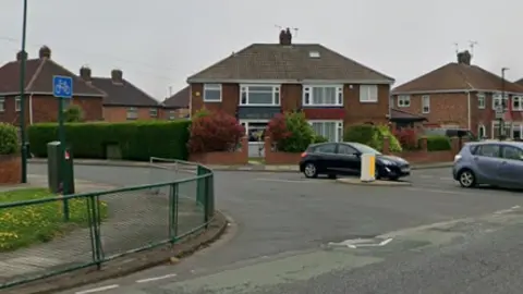 A picture of the junction between Kirkleatham Road and Staintondale Avenue. Metal fencing can be seen on the left side of the road and two cars are waiting to pull out.