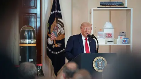 Donald Trump pictured speaking at an event at a podium marked with a circular White House logo. Behind him, there are objects as part of an event highlighting business investment in the US 