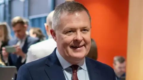 Getty Images A man is looking beyond the camera wearing a navy blue suit jacket, a red tie and a light blue shirt. Behind him are people gathering around, partly in front of an orange wall.