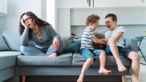 Getty Images Middle age single mother with funny suffering tired face sit on couch in living room together with her fighting little sons
