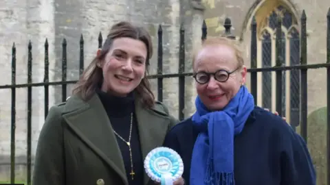 Reform UK A close-up head and shoulders shot of Aimee and Bente smiling to camera and standing in front of railings with a church in the background. Bente is holding a Reform UK rosette in turquoise and white colours