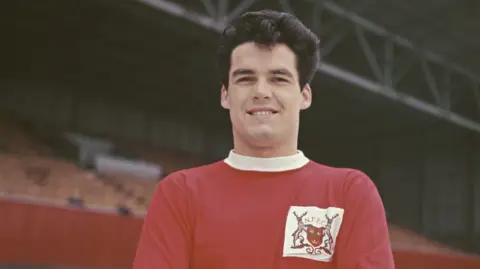 Frank Wignall Nottingham Forest player wearing the 1960's forest strip. A red top with a white collar. The club emblem with the initials NFFC -Nottingham Forest Football Club is displayed prominently. Wignall is stood pitch side in front of one the stands 