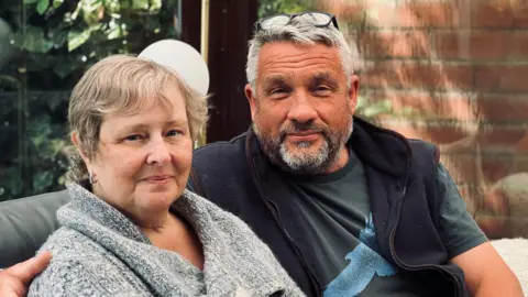 Madeleine Butcher sat with her husband John who has his arm around her and glasses on the top of his head. Madeline is wearing a grey jacket and is looking at the camera. John is wearing a t-shirt with a sleeveless jacket and is also looking at the camera