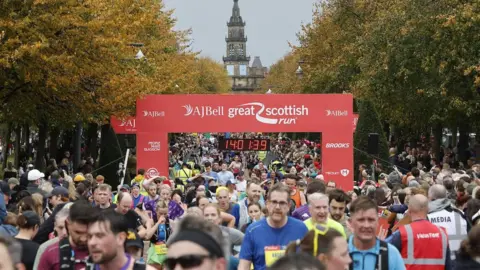 Great Scottish Run The AJ Bell Great Scottish Run, pokazujący dużą grupę biegaczy przekraczających linię pod czerwonym sztandarem. Cyfrowy zegar odczytuje 1: 40: 13.9, a scena zawiera jesienne drzewa i historyczny budynek z wieżą zegarową w tle.