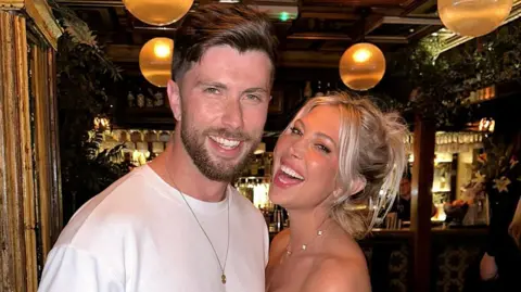 Richie and girlfriend Sarah. They are standing in a bar and both smiling into the camera.