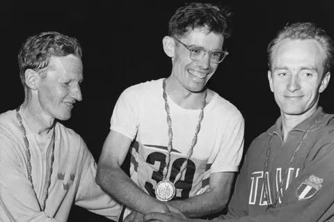Getty Images Three men wearing medals and athletics gear holding hands together in a black and white photograph. All the men are smiling, particularly the man in the centre of the image.