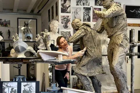 Coastal Forces Heritage Trust Amy Goodman is pictured in her workshop, using a tool on a statue in progress. Near her are figurines of two men on the prow of a boat, showing how part of the Coastal Forces memorial will look. Photographs of Royal Navy Wrens are positioned in front of her.