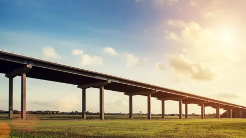 The M62 Ouse Bridge in hazy sunshine. There are green fields in the foreground.