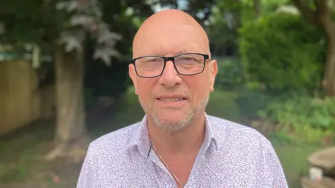 A bald man wearing glasses looks at the camera with a neutral expression