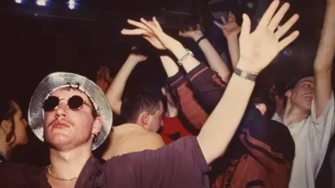 Tony Davis / British Culture Archive  Men and women dancing inside The Eclipse nightclub, with one club-goer sporting a silver hat and sunglasses