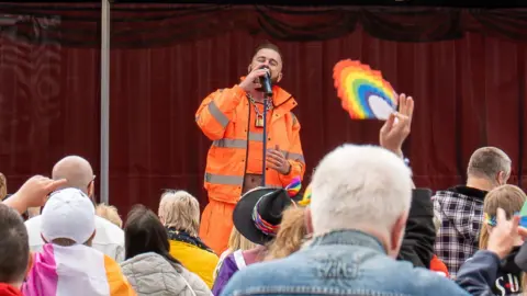 A view of a man in an orange hi vis jacket singing to a crown, with a rainbow being held up by one onlooker.