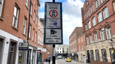 Road sign looking down a city centre street. Sign reads Pedestrian and Cycle Zone and traffic enforcement camera timings