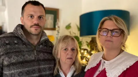 Three people, one man and two women, looking into the camera. The man is wearing a fleece hoodie. The lady in the middle is wearing a white shirt and a grey jumper. The lady on the right is wearing a white blouse, pink fluffy cardigan and a pair of glasses. In the background there is a picture on the wall and a large blue lamp.