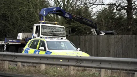 BBC A police car on a road. Behind it is a white and blue recovery vehicle