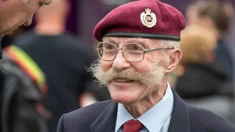An armed services veteran wearing a red beret and spectacles with a wide moustache