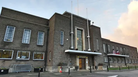 A landscape shot of Havering Town Hall. It is a wide, dark brown brick building with long windows, in an international modern architectural style. 