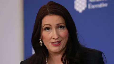 Little-Pengelly has long black hair and is talking in the picture. She is wearing makeup, pearl earrings and necklace. She is standing in front of a blue wall.
