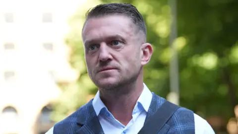 Tommy Robinson is wearing a light blue shirt and a checked blue waistcoat with a messenger bag over his shoulder. He has short hair and light stubble.
