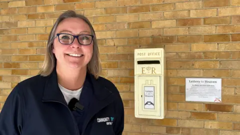 BBC A woman wearing a fleece and glasses standing next to a light golden postbox which says Letters to Heaven on it.