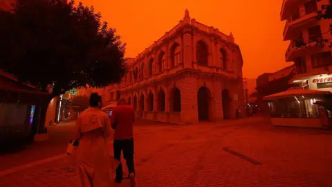 Stefanos Rapanis/Anadolu via Getty Images Two people walk in jackets long a street beside a shop and building. The sky and everything is coloured red-orange.