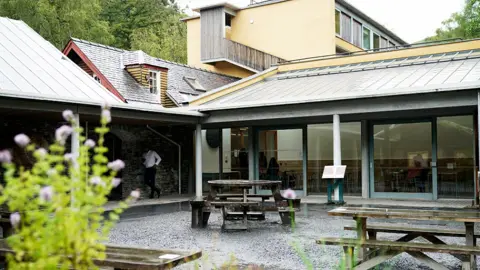 Getty Images In the foreground is a gravel covered courtyard with wooden picnic tables at the Centre for Alternative Technology, Machynlleth. Parts of the building can be seen behind, including an original slate roofed building which has had extensions to the front and both sides adding on more modern, glass fronted buildings with patio sliding doors and solar panels on top. In the background is a flat-topped building with wood-lined balcony.
