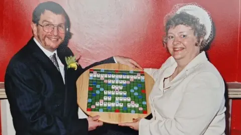 Graham and Helen Harding Graham and Helen on their wedding day holding a scrabble board. Graham is wearing a tux with a yellow rose on his pocket holding a scrabble board with his wife Helen who's wearing a white suite with a hat on her head. 