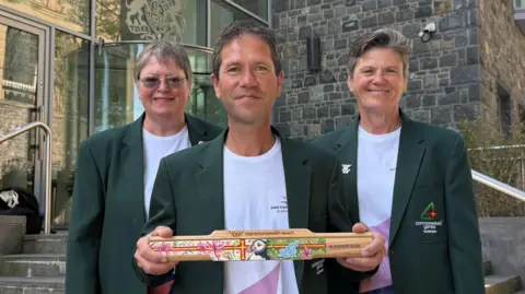 BBC Angela Stuart, Peter Jory and Karina Jackson are wearing green blazers and standing next to one another. They are outdoors in front of a stone building and steps. Peter Jory is in the middle of the group and is holding the baton. 