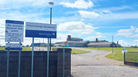 The entrance to Fraserburgh Academy. There is a low wall and three blue and white signs, with one that reads: "Fraserburgh Academy. Aberdeenshire Council". There is a long road from the entrance up to a number of buildings that make up the school.
