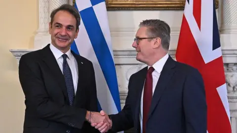 Reuters Starmer and Mitsotakis smile and shake hands at Number 10. Starmer looks at Mitsotakis, while Mitsotakis looks at the camera. The flags of their countries are behind them