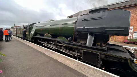 A green steam locomotive is parked by a platform. There are some people standing next to it, one in an orange high-vis vest.