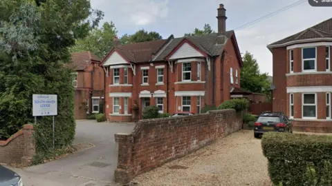 A google street view image of South Haven Lodge Care Home. It is a large red brick building set back from the road with a large driveway. The home's sign is visible at the top of the driveway by the road.