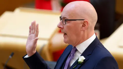 Getty Images John Swinney, who is bald with glasses, speaks with his right hand raised. 