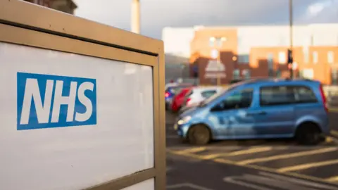 A close up of an NHS sign outside a hospital, with a single car parked in the background.