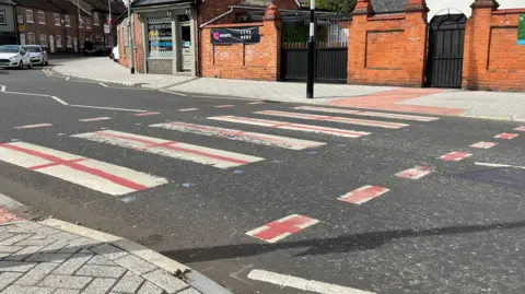 A pedestrian crossing, which is normally a row of white rectangles painted on to the road, with a red painted cross added to each white rectangle.