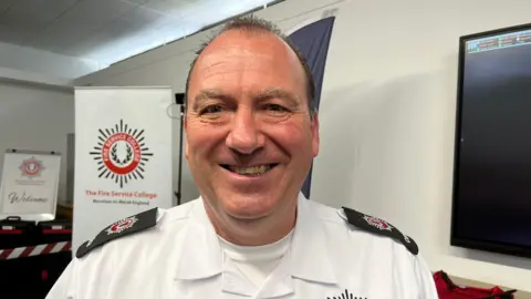 A man wearing emergency service uniform smiles in front of poster saying 'the Fire Service College'.