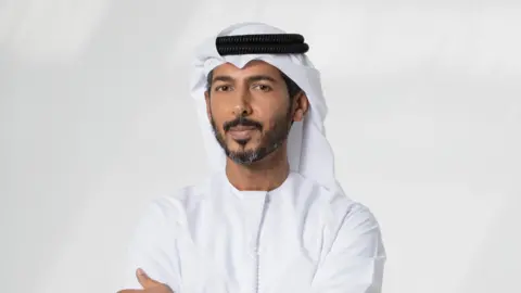 Hassan Alanakabi Hassan cruza o braço e olha para a câmera. Ele está usando os tradicionais Emirados Tihari - um Kandura - um longo vestido branco. Na cabeça, ele usa um sem cabeça branco chamado ghutra e é protegido por um cordão preto chamado Agal.

