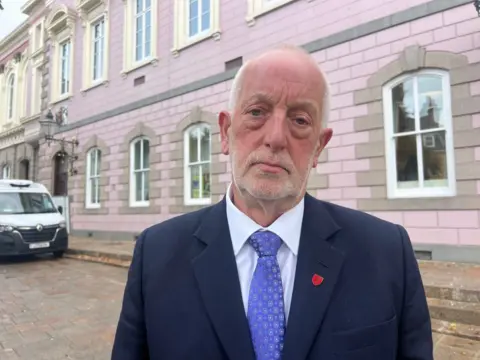 Deputy Steve Luce is stood outside out the States Assembly. He is wearing a dark blue suit and a light blue tie. 