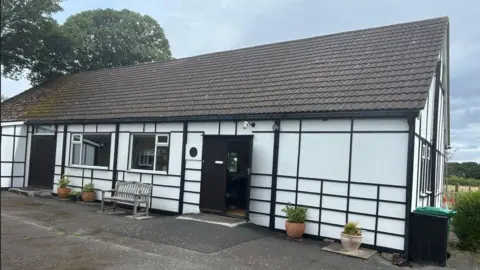 Barnston's Women's Institute is a black-and-white Tudor-style building. There are plant pots lining the front of the building with a bench to the right of the front door.