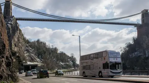 A white double-decker bus travelling under Clifton Suspension Bridge. It is near the River Avon and there are cars either side of the carriageway. 