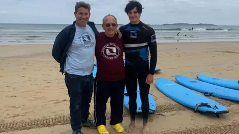 Andy, Ian and Ethan with their arms around each other smiling at the camera with the sea and surf boards in shot behind them lying on the sand. 