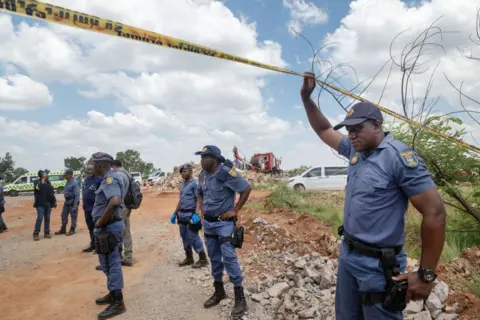 Getty Images Police officers stand on a dirt road as one of them in the foreground raises his arm to hold aloft some police tape.