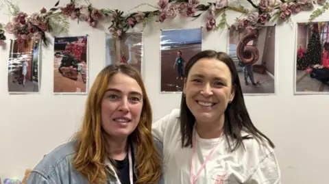 Two women looking into the camera smiling. One has ginger hair and the other has dark brown hair. Behind them are different photographs of her daughter, Jessie, who is fighting cancer in hospital.