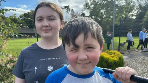 BBC Ellie and Zack standing at the miniature golf course. Ellie is wearing a grey t-shirt and has her dark hair pulled back in a ponytail. Zack has short dark hair and is wearing a blue and grey top. He is squinting because of the sun and is carrying a putter in one hand