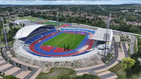 An aerial view of the stadium, which dominates the photo. Covered stands by the blue athletics track are on the left and right of the image. Trees and houses are in the background.