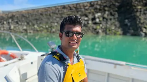 Shetty Chavan wears dark sunglasses, a grey shirt, and a bright yellow waterproof bib, sitting on a small boat. The boat is on turquoise water, with a stone harbour wall in the background. He's turned slightly to one side, facing the camera, and sunlight creates strong highlights and shadows across the scene.